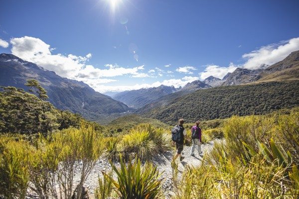 two people walking on trail under blue skies and sun with winding bush clad valley in background